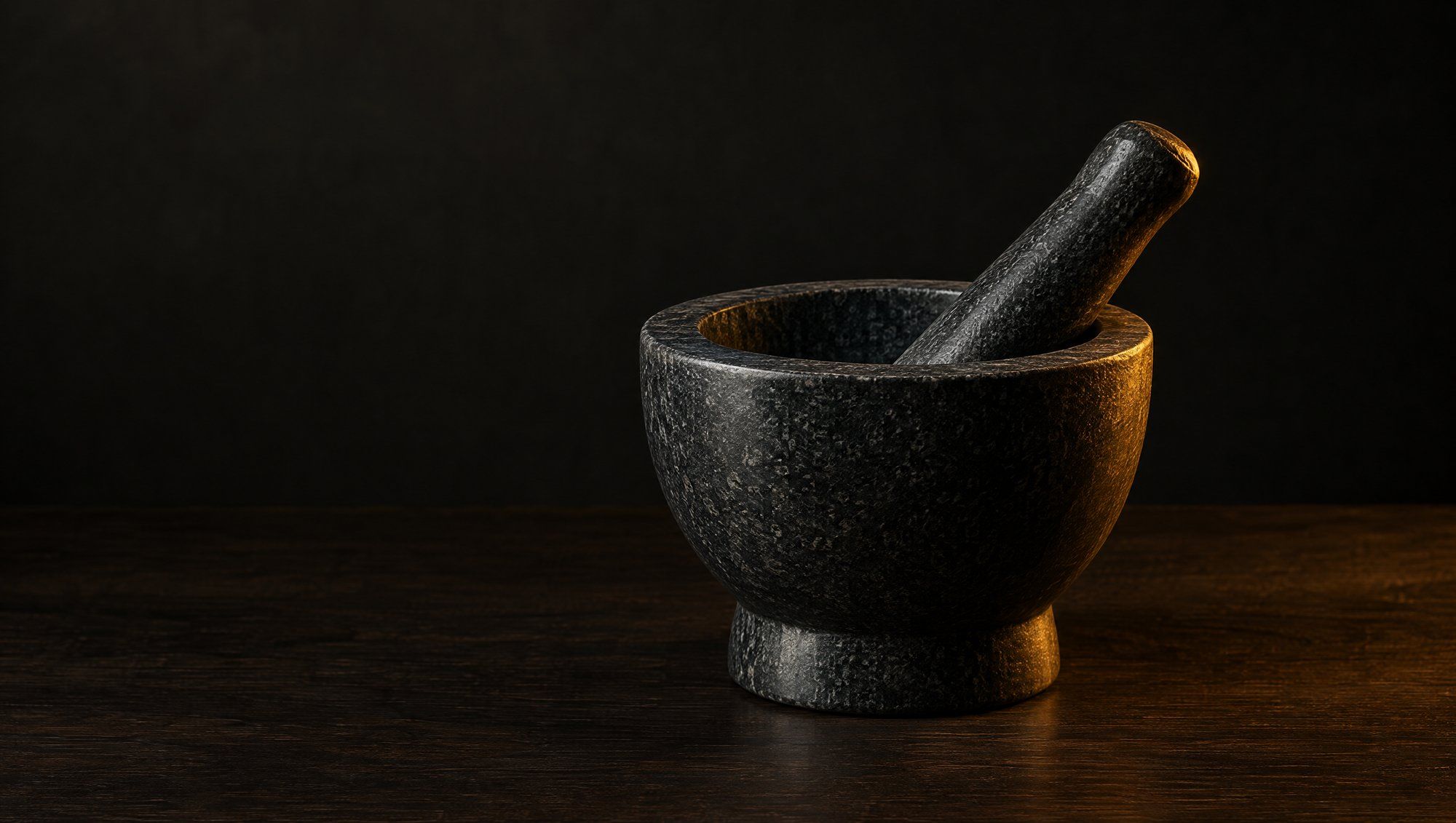 An empty dark granite mortar and pestle on a wooden counter in low warm light